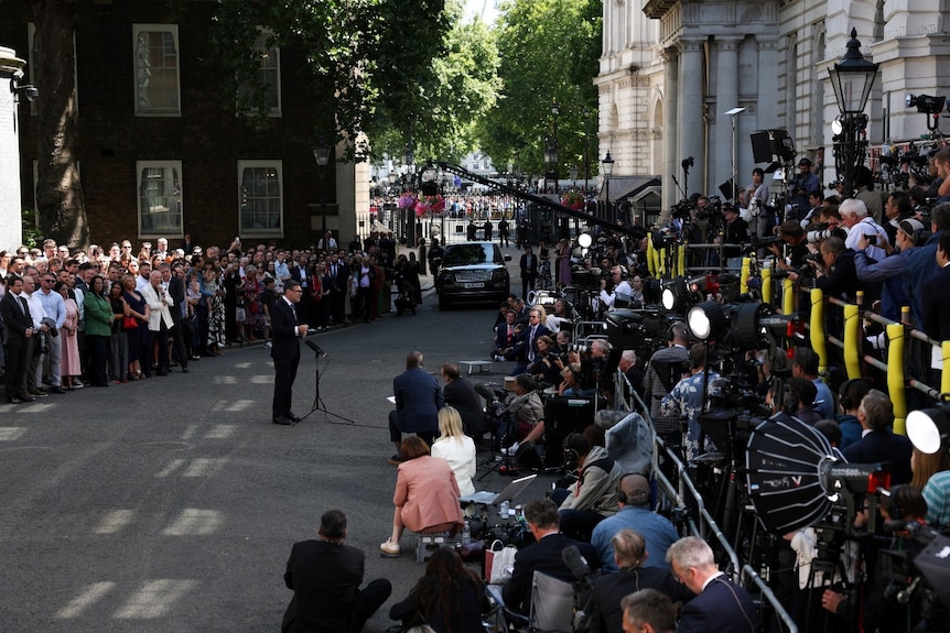 A man in a suit speaking on a street. Large crowds are visible around him.