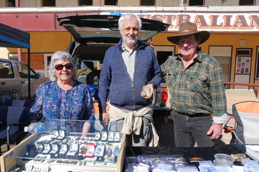 Three smiling older people behind a stall at an open-air market.