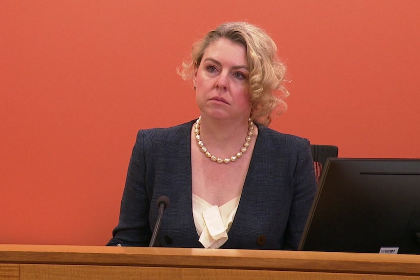 A woman wearing a suit sits behind a large wooden bench.