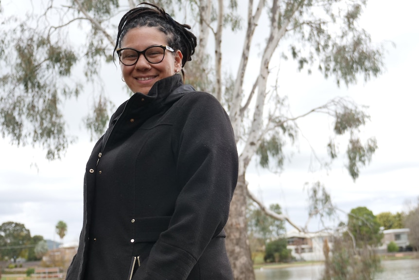 A woman in a black coat smiles at the camera in front of a tree
