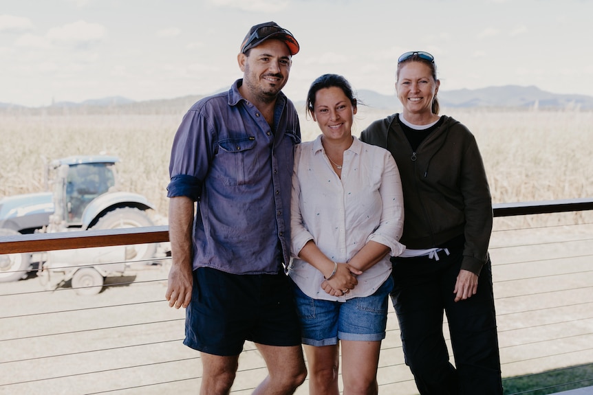 Three people lean on a veranda railing with a tractor and farmland behind them