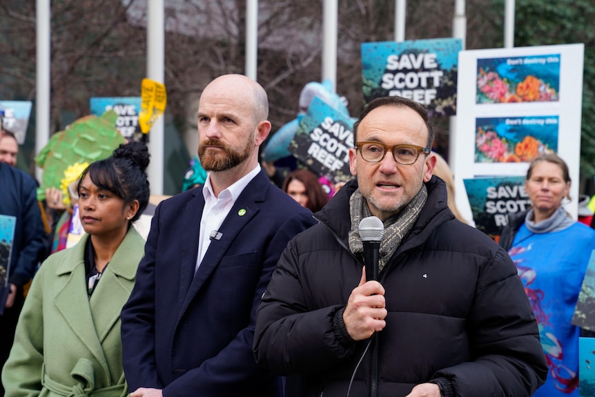 A man in glasses speaks into a microphone while demonstrators hold signs reading "Save Scott Reef" behind him.