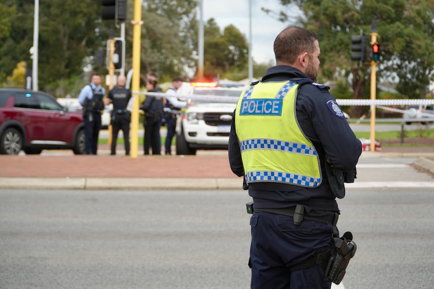 A police officer in hi-vis stands in front of other officers and a police vehicle at a crime scene at an intersection.