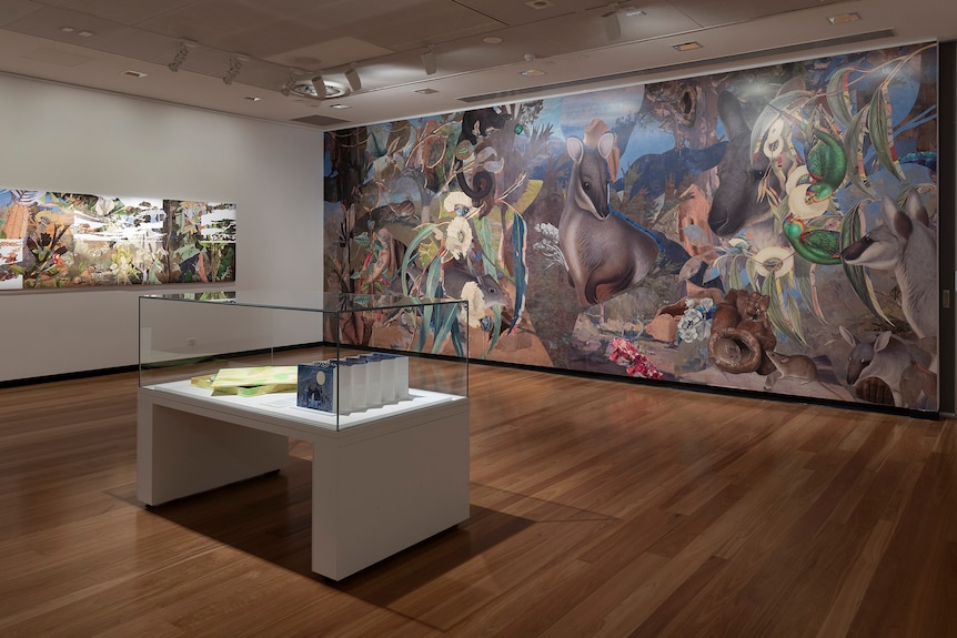 An artwork depicting Australian native flora and fauna on the wall of a gallery; a glass cabinet in the middle of the room.