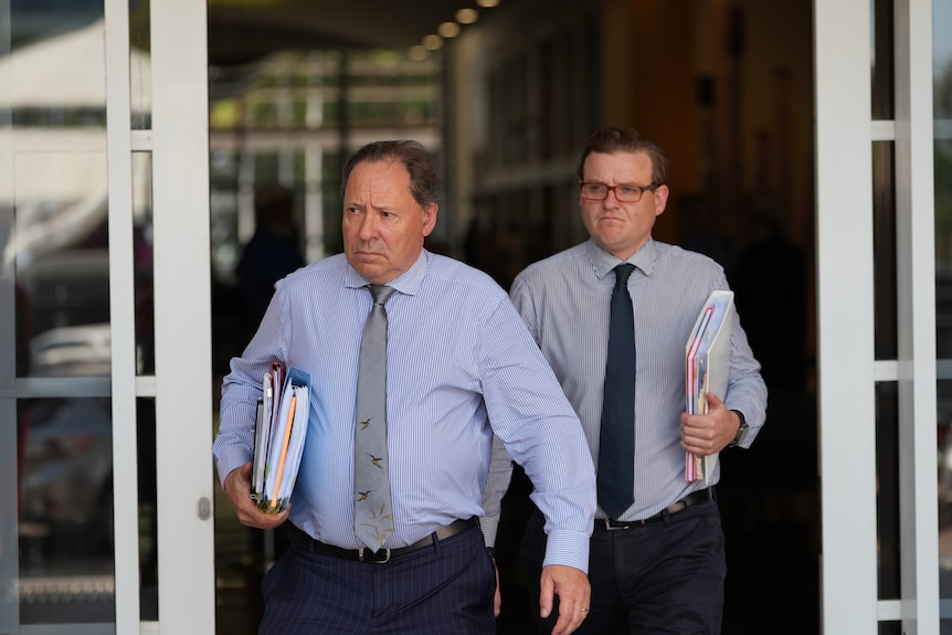 Two white men wearing button up shirts and ties, dark business pants, holding folders and walking out of court.