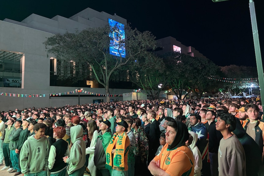 Soccer fans watch intently at a night-time viewing party with a city building behind