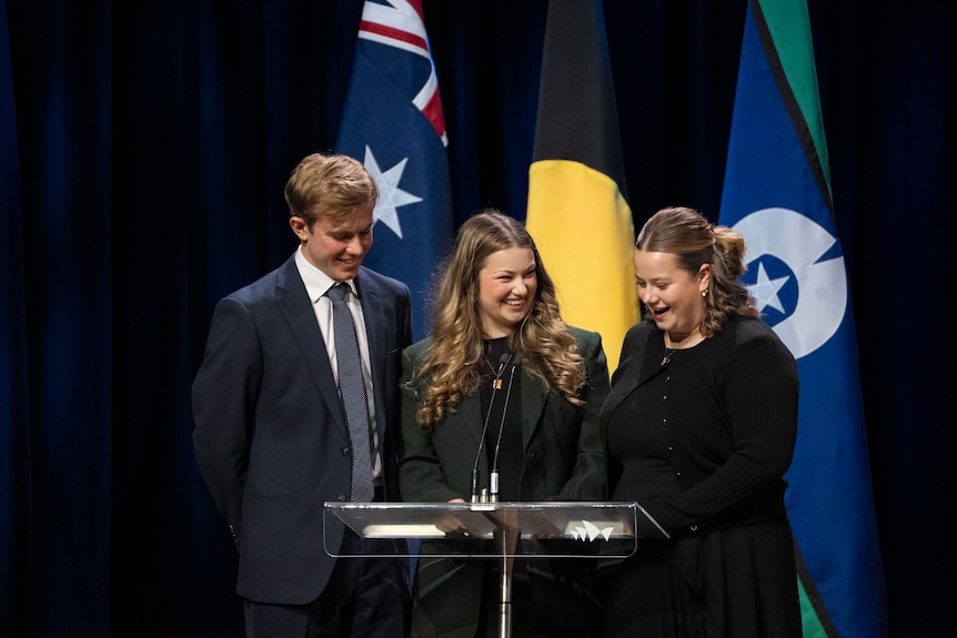 A man and his two sisters standing at a podium in front of flags.