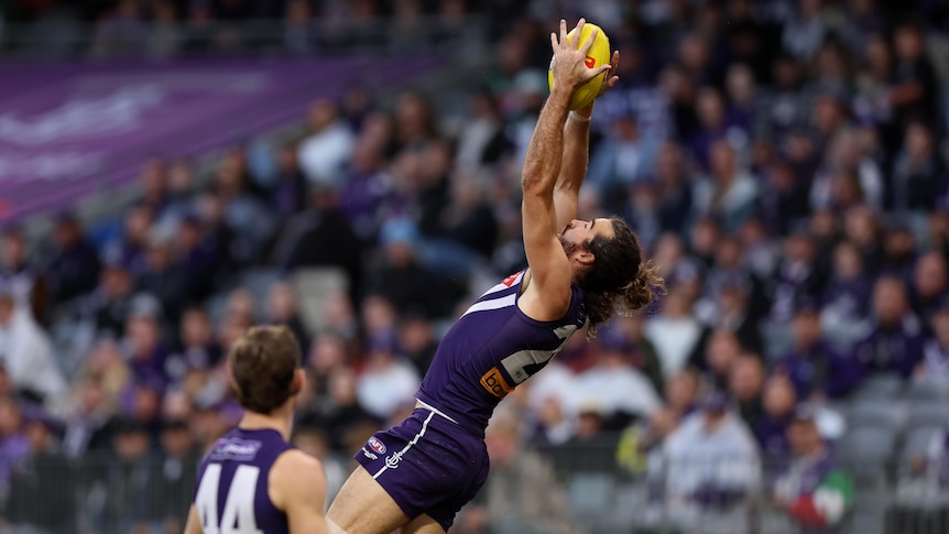 A Fremantle AFL player stretches his arms out above his head to take an athletic mark as a teammate stands by. 