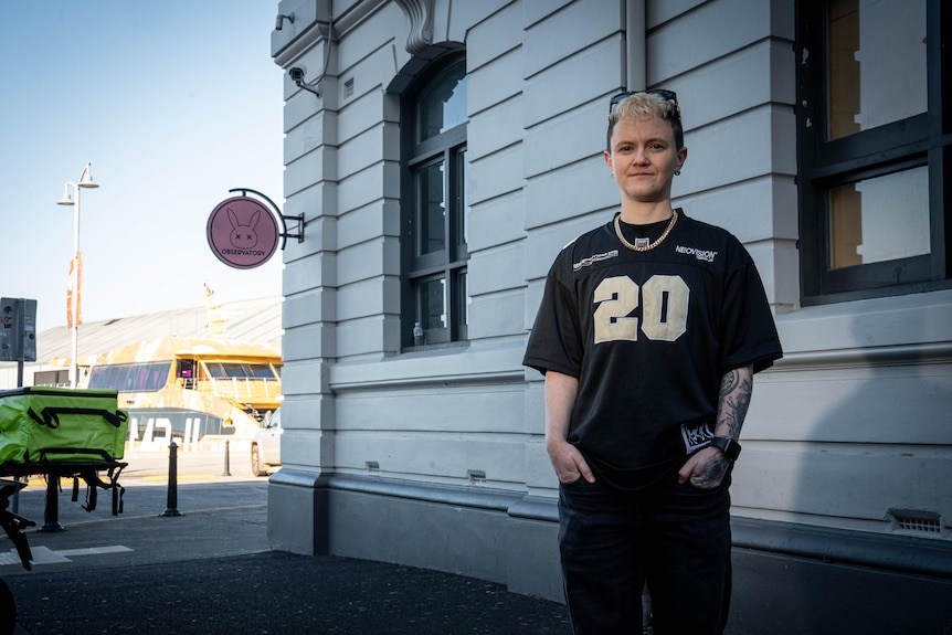 A female with a blonde pixie haircut in a black tee and pants stands outside the Observatory Bar, hands in her pockets.