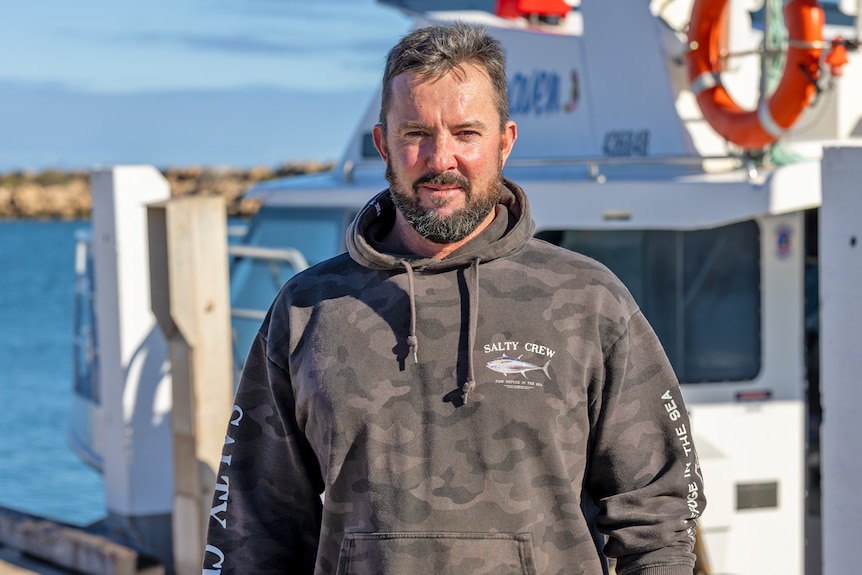 Jonathan is wearing a brown and grey hooded jumper standing in front of a cray boat.