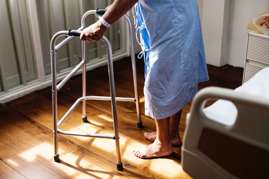 A person in a pale blue hospital gown holds onto a walking frame.