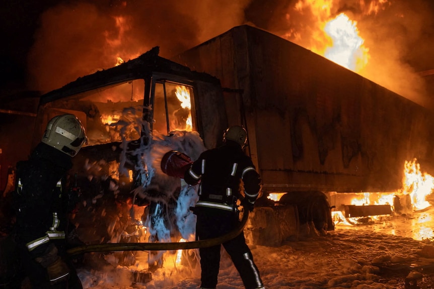 Two firefighters silhouetted against a truck engulfed in flames.