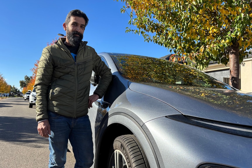 A dark-haired, bearded man in a jacket stands near a car parked on a suburban street.