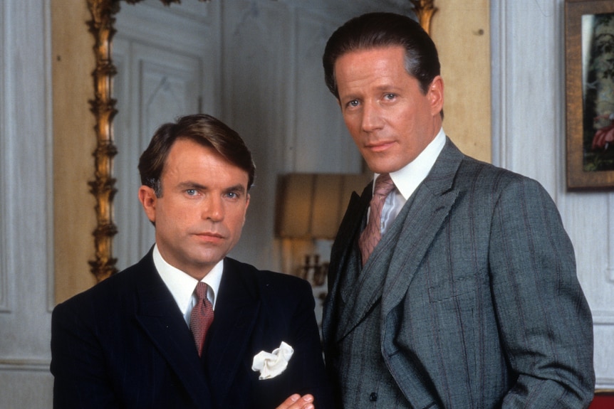 Two men in sharp suits and ties look serious. One man seated has his arms crossed while the other stands with hands in pockets 