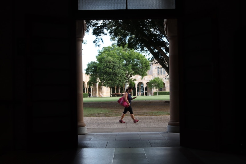a student walks past a large sandstone archway at a university campus looking at their phone
