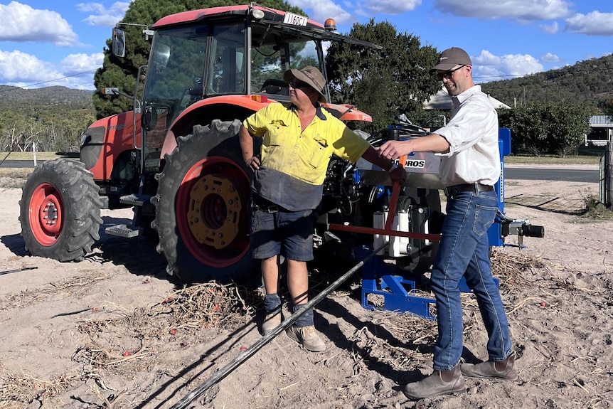 Two men stand near a tractor in a paddock as they lay out a long strip of black plastic.