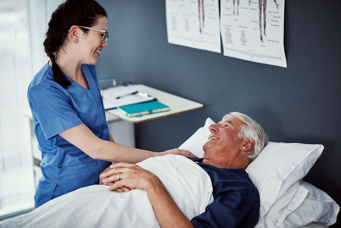 A nurse smiling at an elderly male patient in a hospital bed, discussing uncomfortable truths about the medical industry.