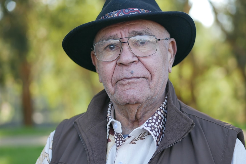 A middle-aged man in a dark hat and a vest stands in a park.