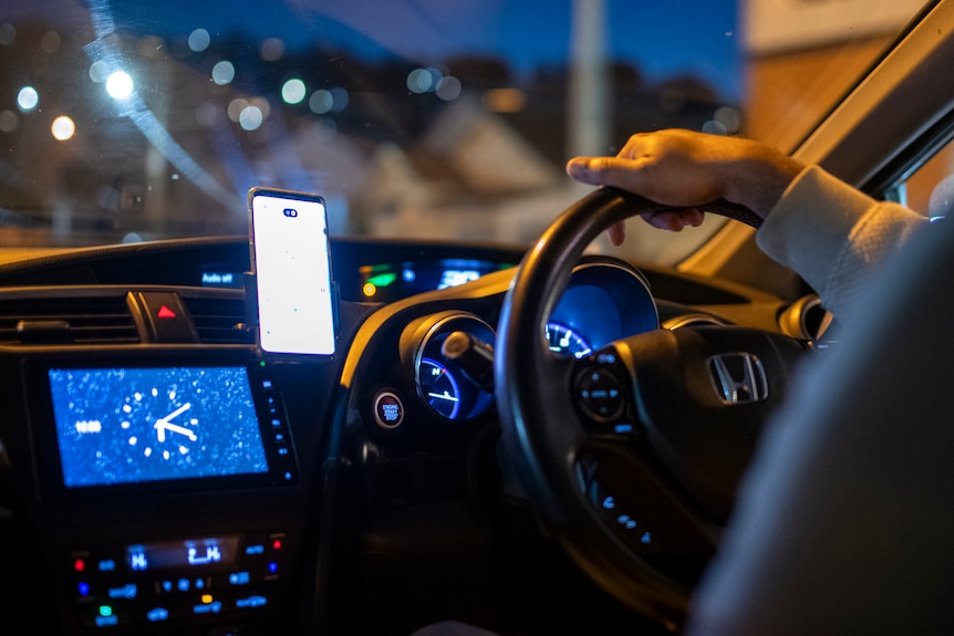 Gold street lamps illuminate a hand on a steering wheel, photographed from inside a car at dusk.
