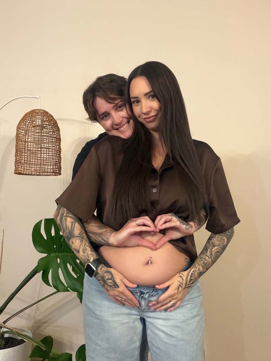 A woman standing behind her female partner making a heart shape with her hands above her partner's belly.
