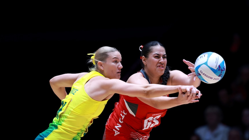 Two netballers grimace as they compete for the ball, an Australian defender trying to punch it and a Tongan trying to grab it.