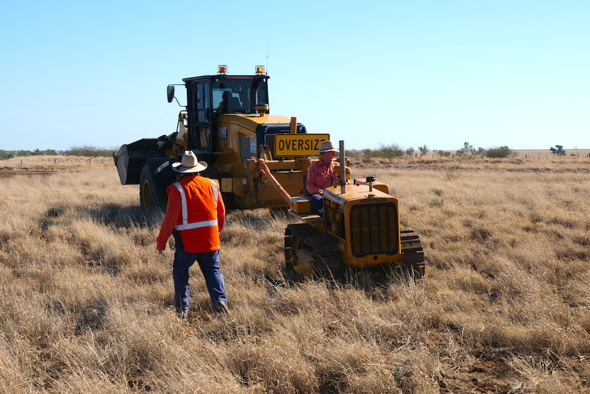 a man rides on a small tractor backwards through long grass, pulled by a larger tractor