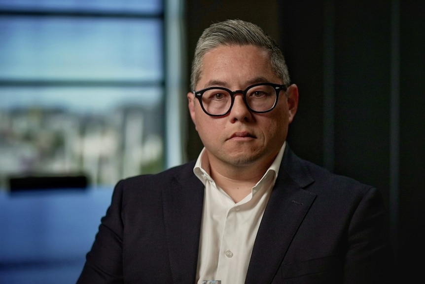 Man wearing glasses, blazer and open neck shirt stares at the camera