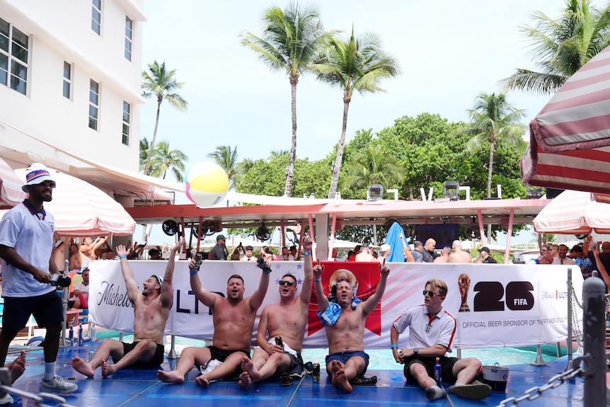 Five men, four with shirts off, sit with their arms raised in the air, cheering.