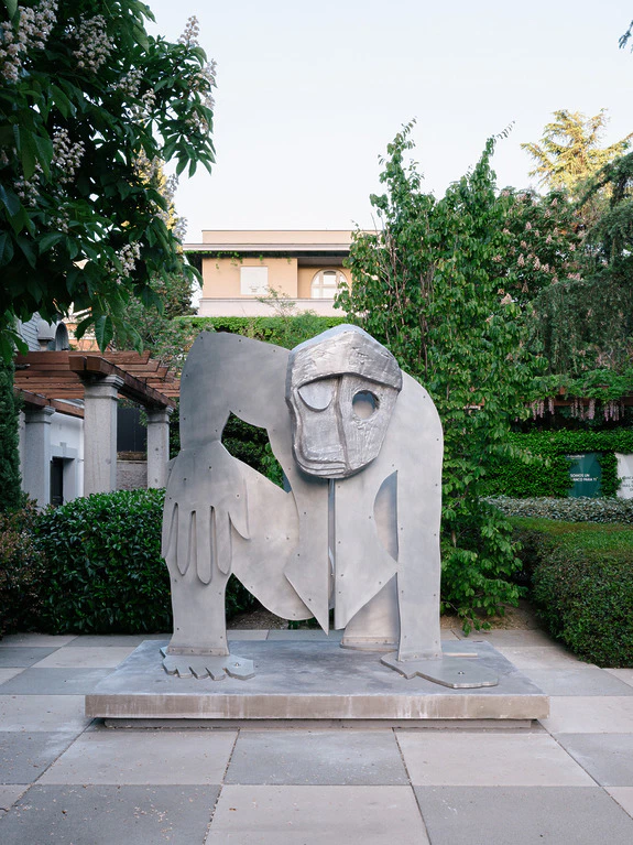 Sculpture of a crouching figure with a mask-like face, surrounded by greenery in an outdoor garden setting.