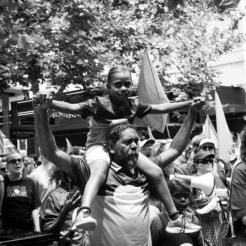A black-and-white photo of a smiling Aboriginal girl on her dad's shoulders as they march through the city.