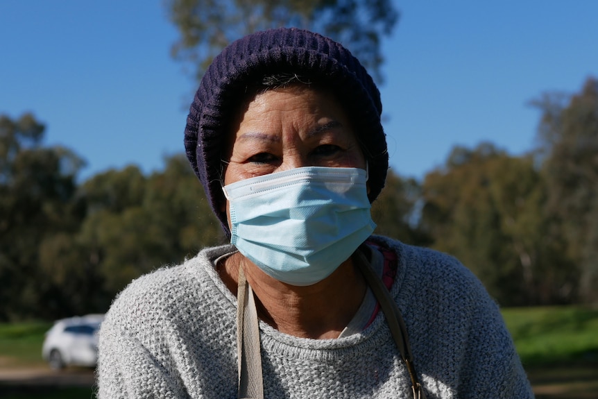 A woman wearing a surgical mask and a beanie. She stands in a park.