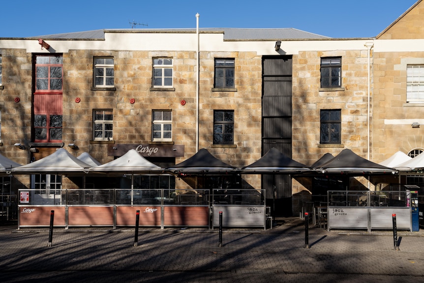 Tall sandstone buildings along a quiet road with umbrella-covered seating out the front and brown fencing.