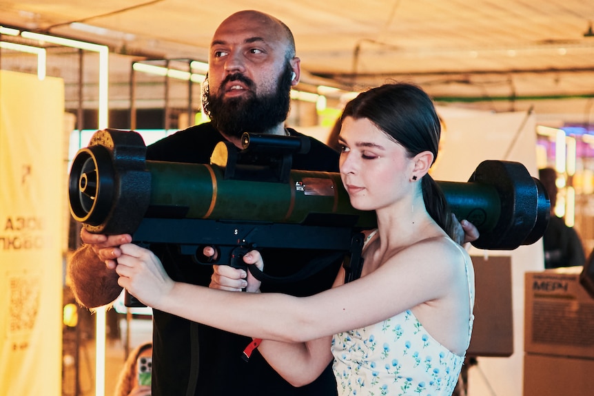 A young woman holds what appears to be a rocket launcher over her shoulder.