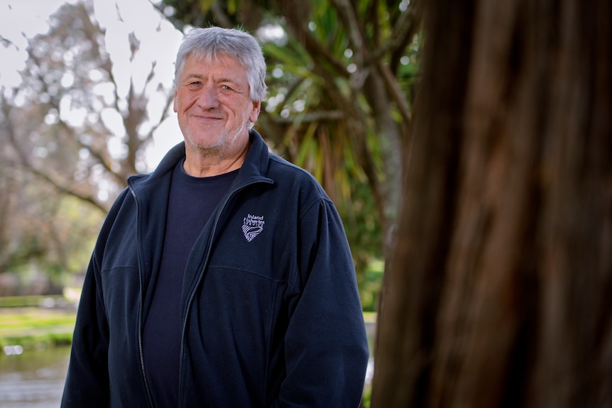 Rob Freeman, a man with grey hair wearing a blue Inland Fisheries jacket stands beside a tree, smiling