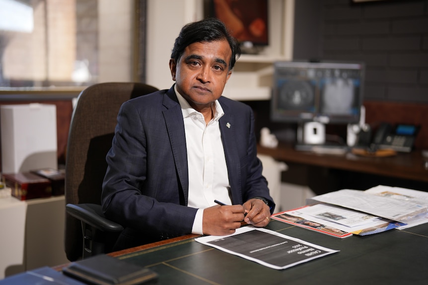 Peter Subramaniam is wearing a suit as he sits at a desk while holding a pen
