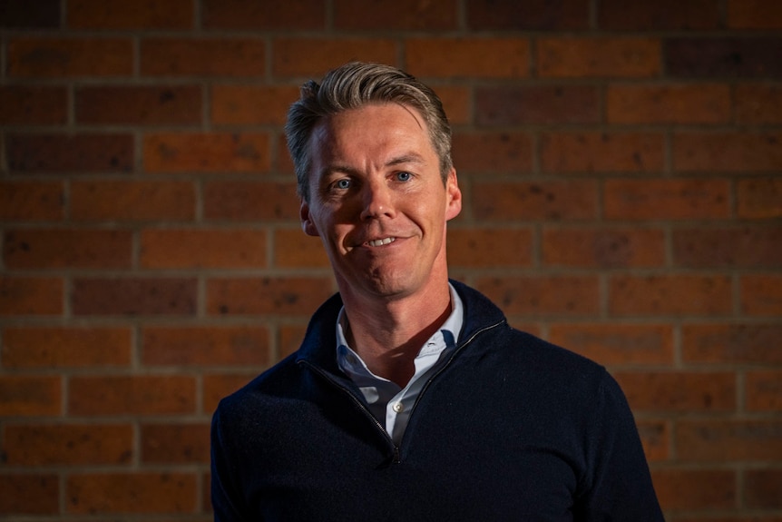 a man with grey hair dark top inside with a brick wall behind him