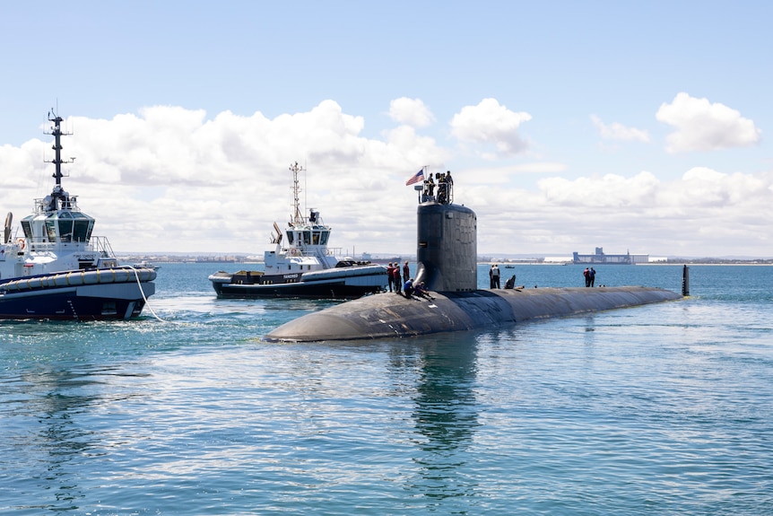 A virginia-class submarine above water, with naval officers standing on top. Two boat ships are nearby.