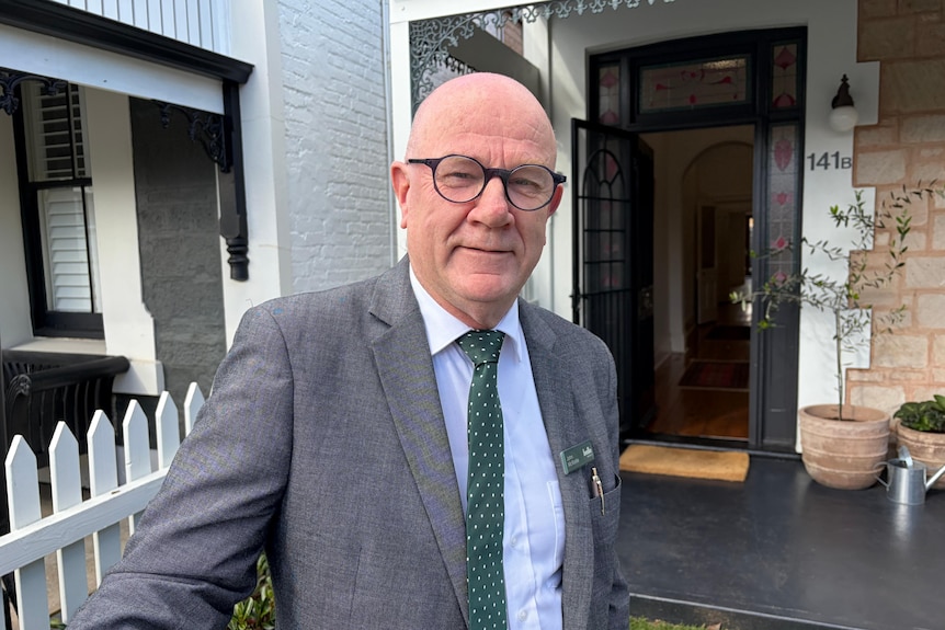 A bald man with black horn-rimmed glasses, grey suit and green tie, stands in front of an open front door to a terraced house.