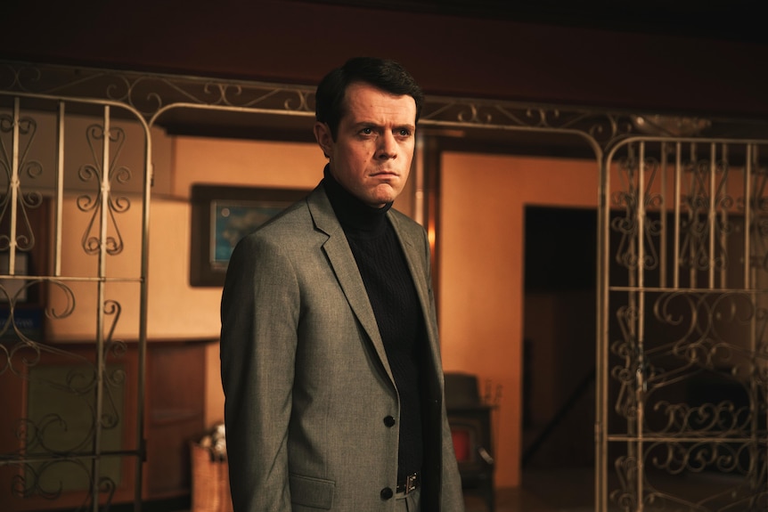 Paul Williams in a 70s style room looking very serious in a black turtleneck and brown suit jacket