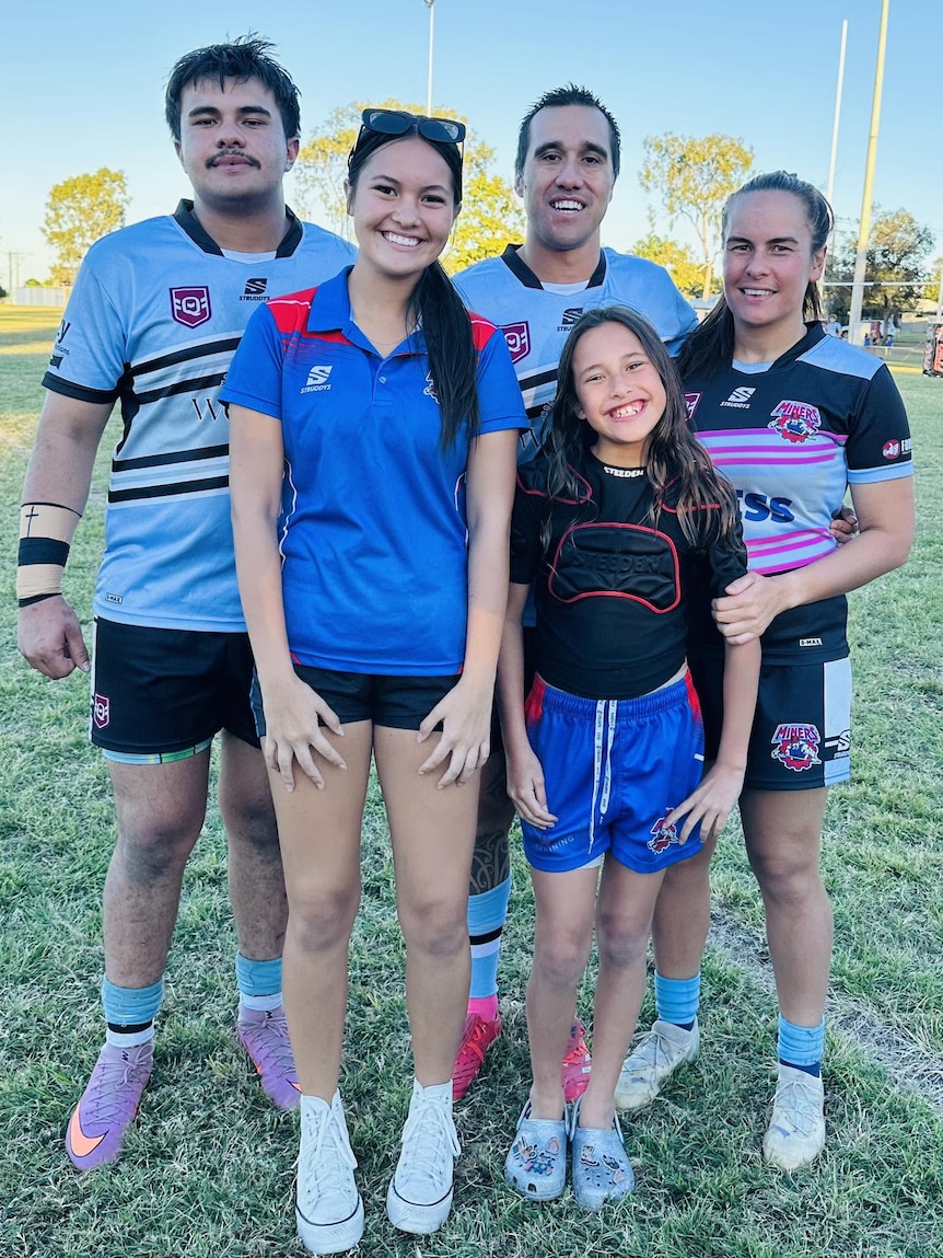 Young family, mum dad and three kids all in same footy shirts and uniforms, smiling at camera