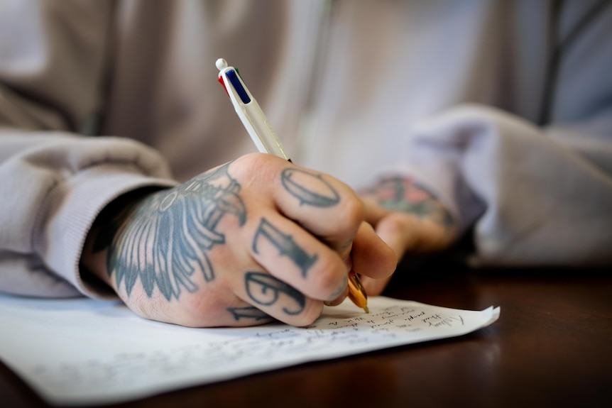 Kieran's hand as he writes on white paper, resting on a brown table. He wears a purple hoodie and his hand has tattoos.