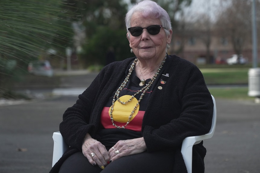 Aunty Brenda Hodge, wearing red white and yellow, as well as shell necklaces, sits in an outdoor courtyard,