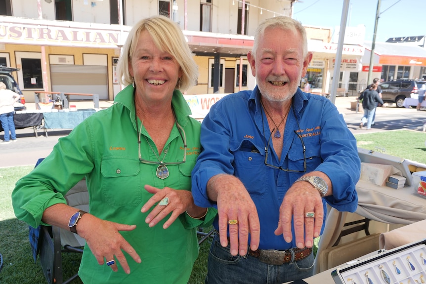 A smiling blonde woman and an older, bearded man smile while showing off their rings at an open-air stall.