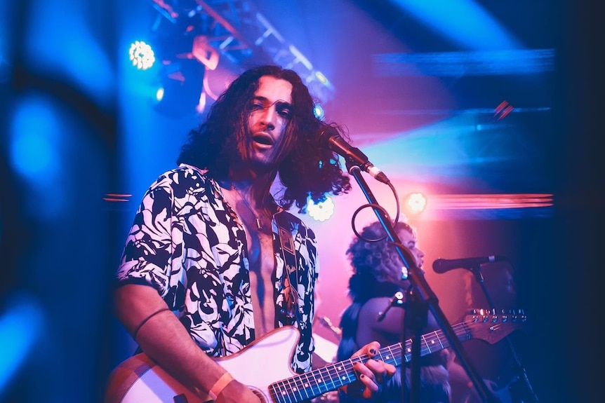 A man plays electronic guitar on a stage with neon lights around.