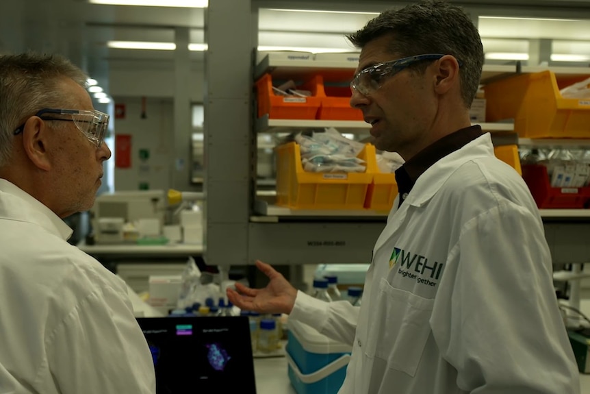 Two men wearing lab coats inside a laboratory, one talks, the other listens.