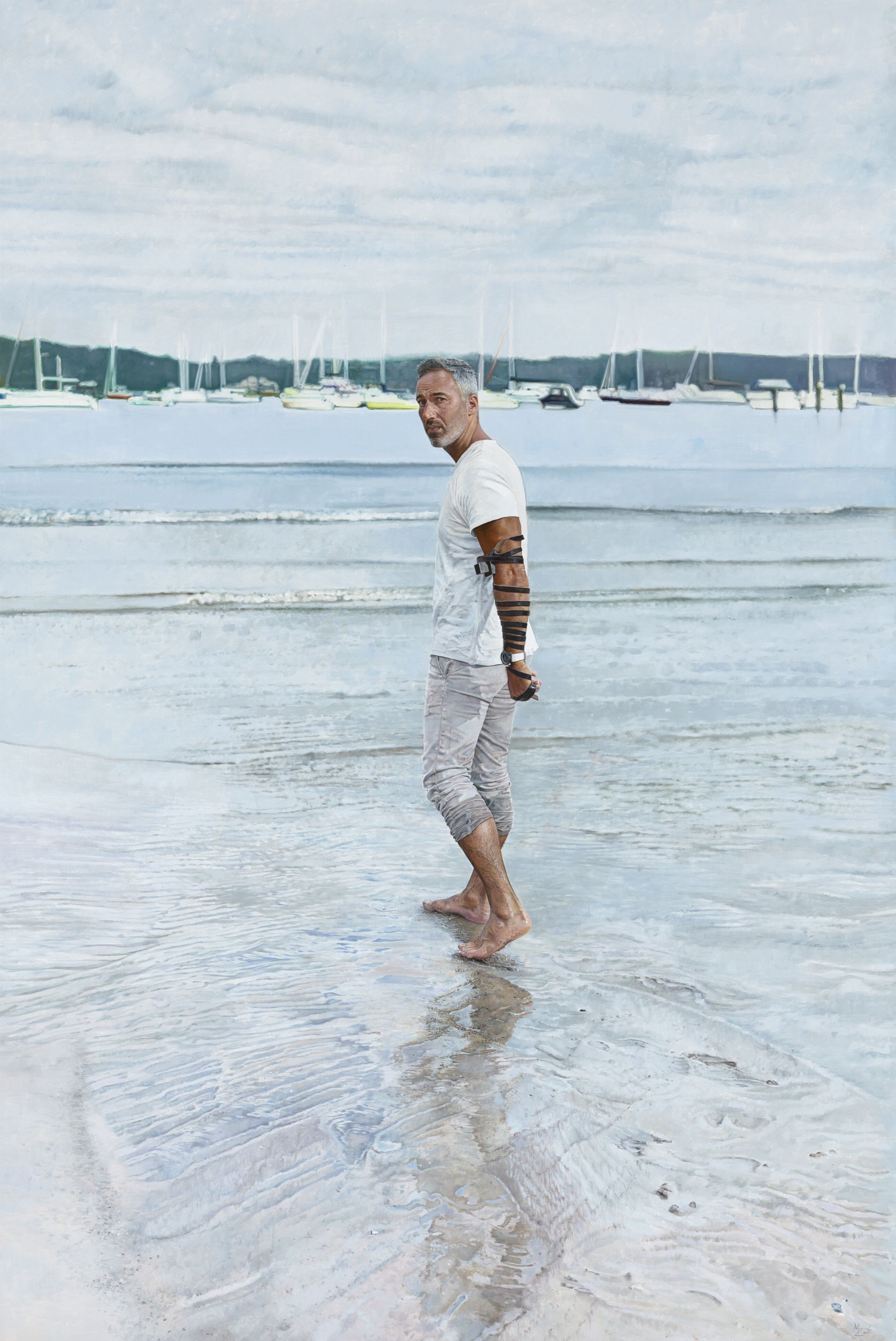 A portrait of a man in a white t-shirt standing at the edge of the water on the beach, boats in the background
