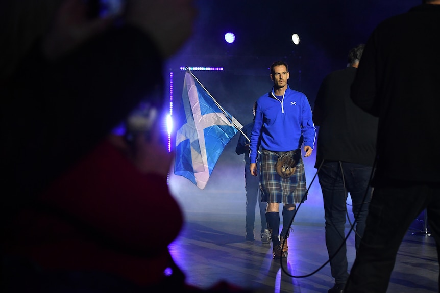 Andy Murray walks in a kilt with a Scotland flag behind him