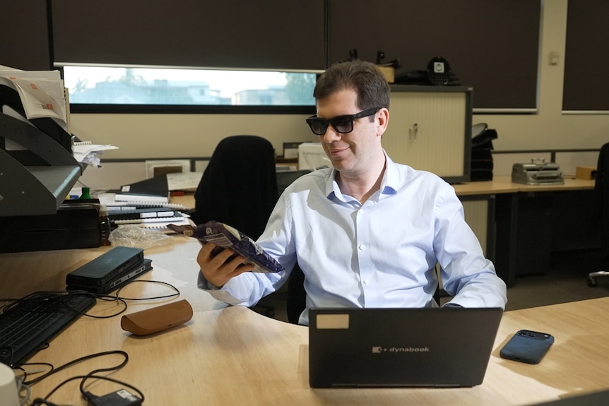 Sam sits at his desk holding a packet of chocolates wearing his glasses and a blue business shirt.