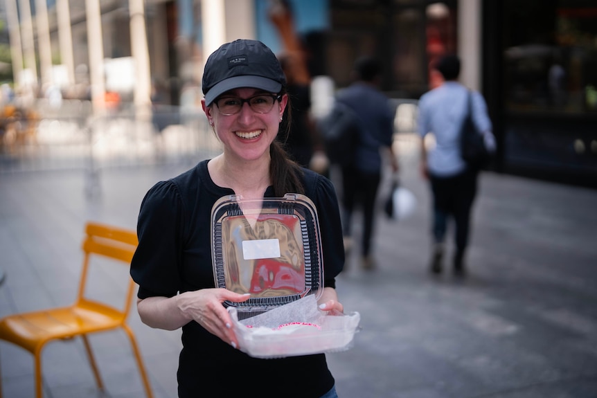 A woman wearing glasses and a black cap holds a takeaway container outside MSG.