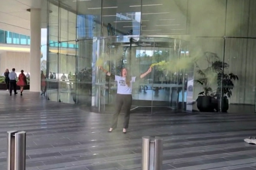 A woman stands outside an office building, releasing green gas from vessels in each hand.